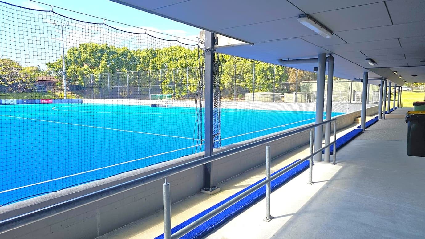 A blue artificial turf sports field is visible through a netted barrier on the left. The field is surrounded by trees and fencing. On the right, there is a covered walkway with a concrete floor and metal railings. The ceiling is lined with fluorescent lights. A row of metal poles supports the structure, and there are bins placed along the walkway.