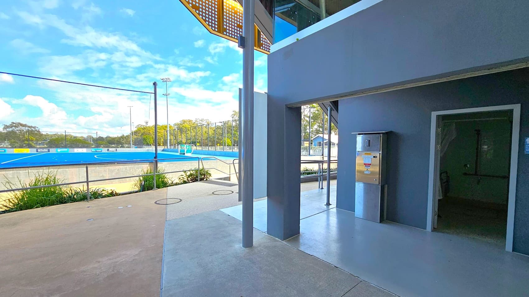 A blue sports field is visible on the left, surrounded by a fence and greenery. In the middle, a concrete path leads to a building with a dark exterior. On the right, there's an entrance to a room with a metal vending machine or dispenser nearby. The ceiling has a perforated metal design, and the floor is a mix of concrete and smooth surfaces. Trees and floodlights are in the background.