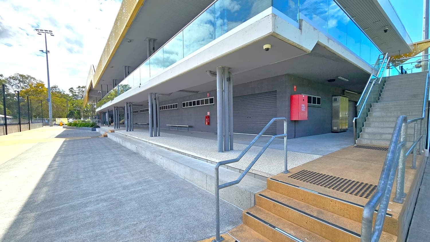 A modern building with a glass facade on the upper level, supported by grey pillars. On the right, a staircase with metal railings leads to the upper floor. Below, a closed roller door and a red fire hose reel are visible. The ground is a mix of concrete and textured surfaces. To the left, a wide walkway extends alongside the building, with a tall light pole and trees in the background.