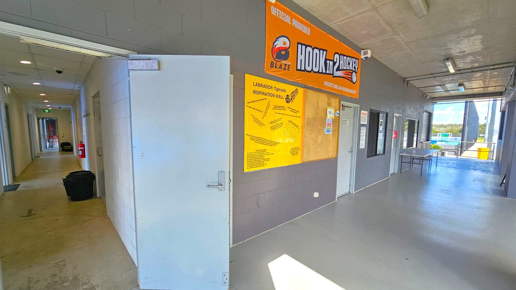 A corridor with a grey concrete floor and ceiling. On the left, an open door leads to a hallway with a fire extinguisher and a bin. On the right, a grey wall features a yellow "Inspiration Wall" board and a large orange banner above. Several doors and windows are along the wall, with a table nearby. In the background, an open area with a view of a sports field and a yellow bin is visible.