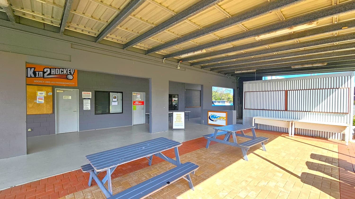 A covered outdoor area with a corrugated metal ceiling and exposed beams. On the left, there is a grey wall with a sign reading "K in 2 Hockey" above a door labelled "Meeting Room 1". Adjacent is a window with notices. In the centre, a counter with a sign and a closed shutter. On the right, a corrugated metal wall with a bench. The floor is a mix of grey concrete and red brick. Two blue picnic tables are in the foreground on the brick section.