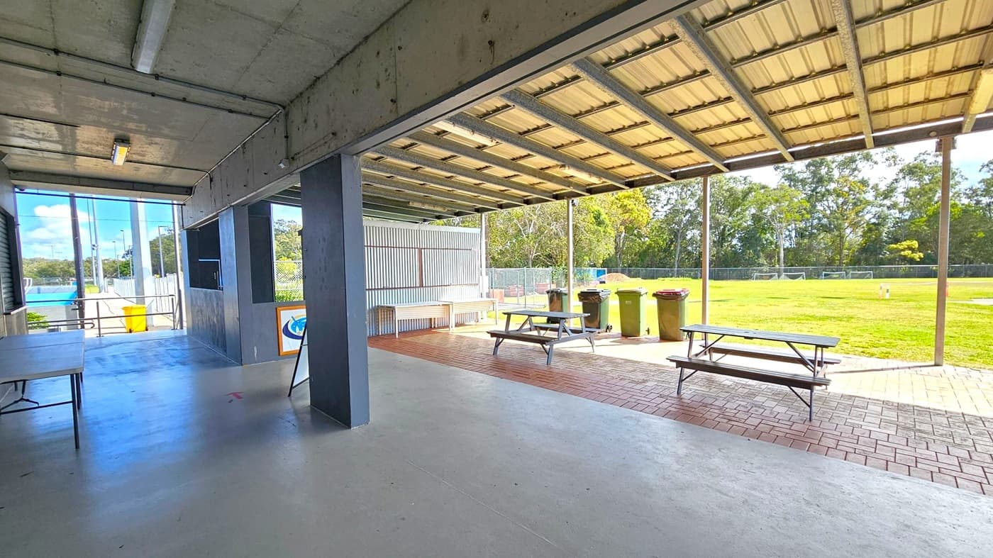 On the left, there is a concrete floor with a folding table and a yellow bin near a railing. The ceiling is metal with fluorescent lights. In the middle, a dark pillar supports the structure. On the right, there are picnic tables and several bins on a paved area. Beyond, a grassy field with trees and small goalposts is visible. The area is partially covered by a metal roof.