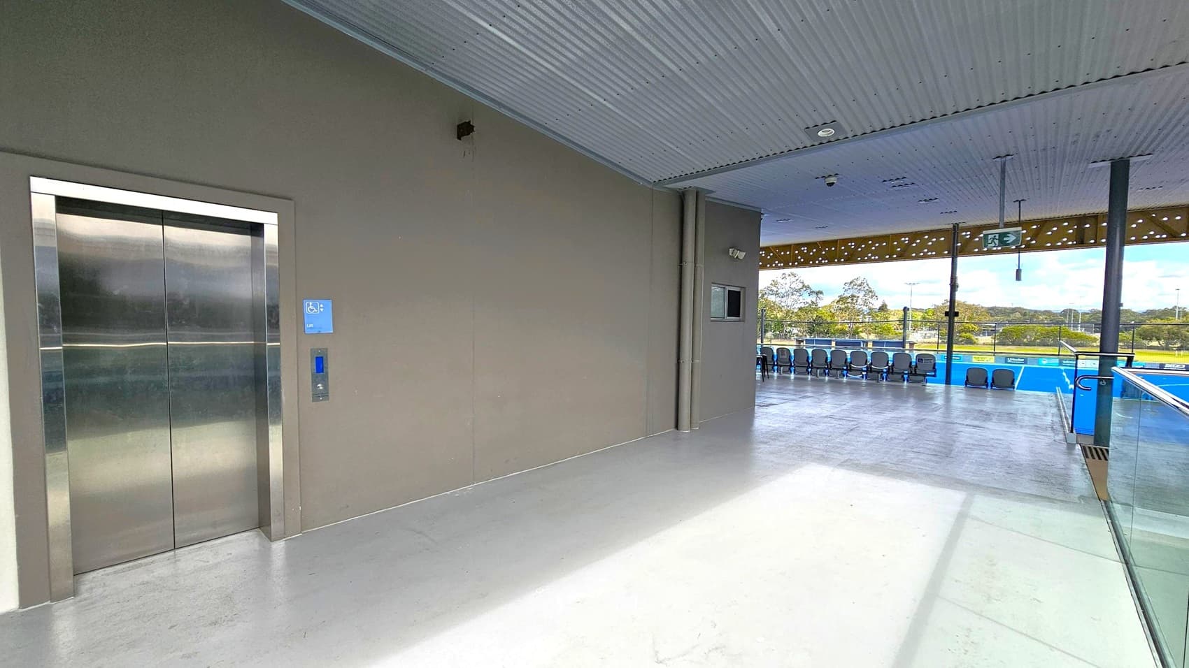 On the left, there is a metallic lift with a blue accessibility sign. The floor is smooth and grey. The ceiling is ribbed and grey. On the right, there is an open area with a row of blue chairs facing a sports field. The area is enclosed by glass railings, and there is an exit sign above. The background shows a grassy field with trees and a partly cloudy sky.