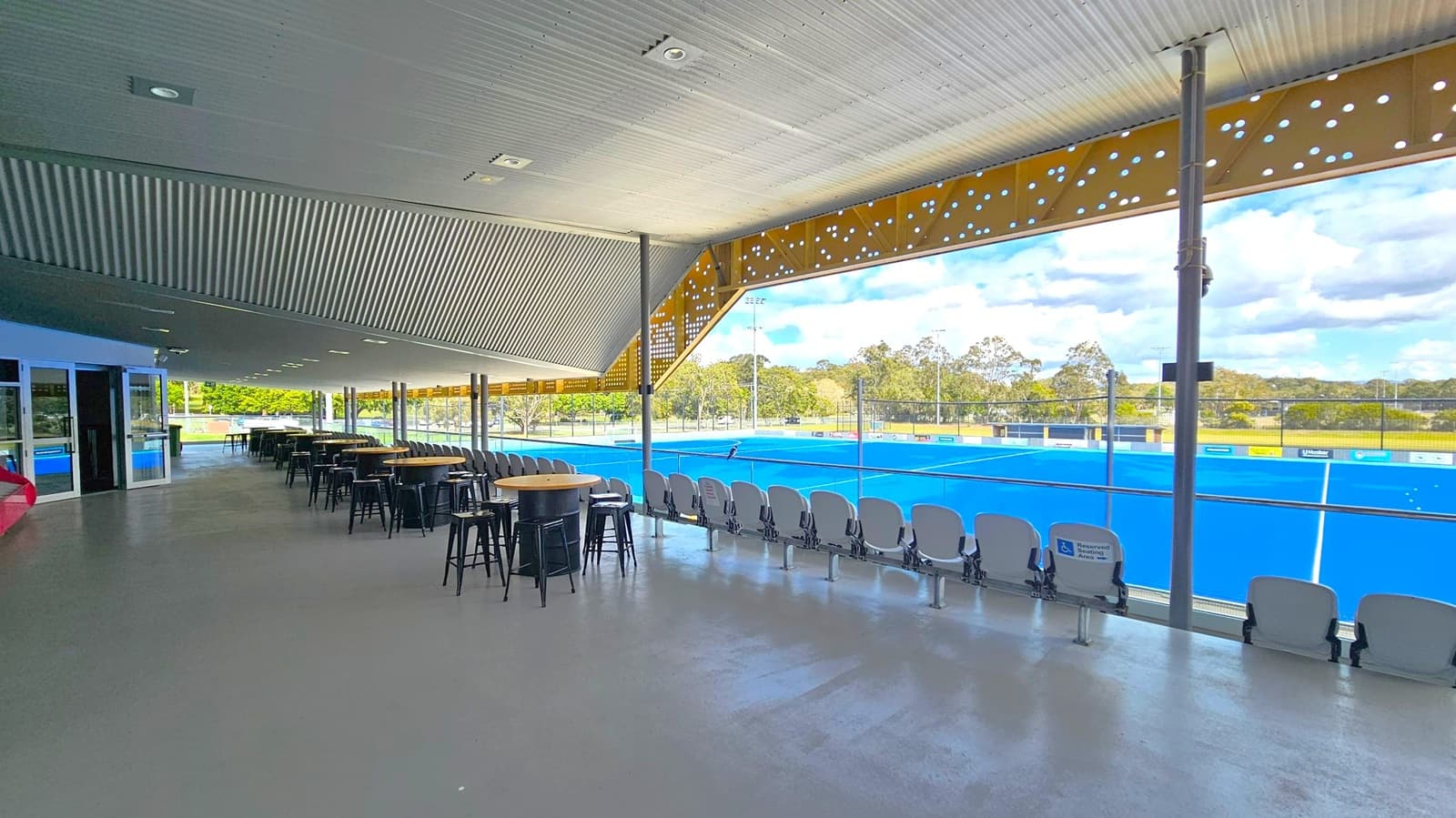 A spacious indoor viewing area with a slanted, ribbed ceiling and a polished concrete floor. On the left, there are tables and high stools arranged in rows. The right side features a row of stadium-style seats facing a bright blue sports field visible through large glass windows. The background shows a field with trees and a partly cloudy sky. The ceiling has recessed lighting, and the area is supported by metal columns.