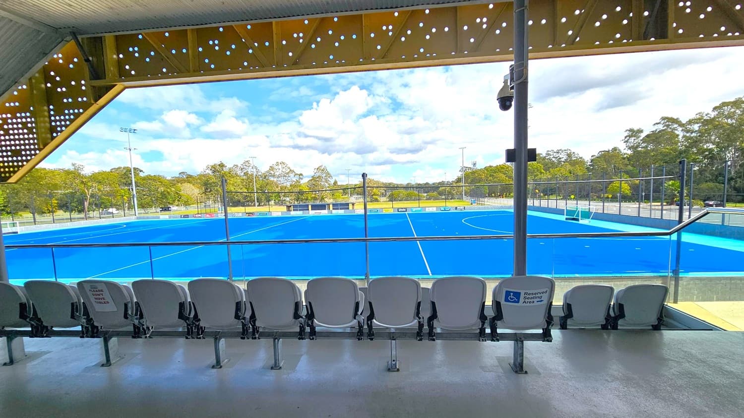 A row of white stadium seats is in the foreground, with one marked as a reserved seating area. Beyond the seats, a bright blue sports field is visible, surrounded by a fence. Trees and greenery are in the background. The ceiling above has a perforated design with circular holes, and a security camera is mounted on a pole. The floor is smooth and light-coloured.