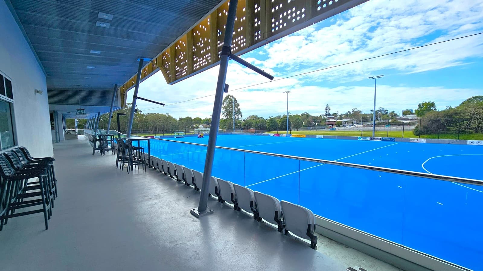A covered viewing area with a grey concrete floor and a perforated metal ceiling. On the left, there are stacked chairs and tables along the wall. The right side features a row of stadium seats facing a bright blue sports field, separated by a glass barrier. The field is surrounded by trees and a few buildings in the background. The area is supported by angled metal beams.