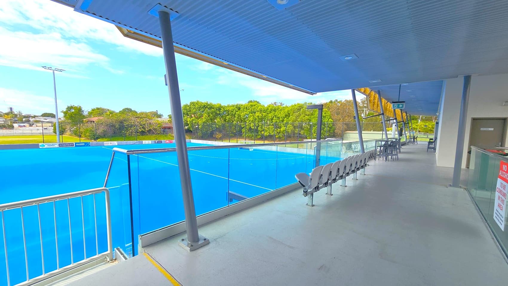 A sports facility with a bright blue playing field on the left, surrounded by a glass barrier. On the right, there is a row of white seats and a few tables with chairs under a blue corrugated metal roof. A sign on the wall reads "No alcohol to be consumed in this area." In the background, there are trees and a few buildings. The floor is a light grey concrete.