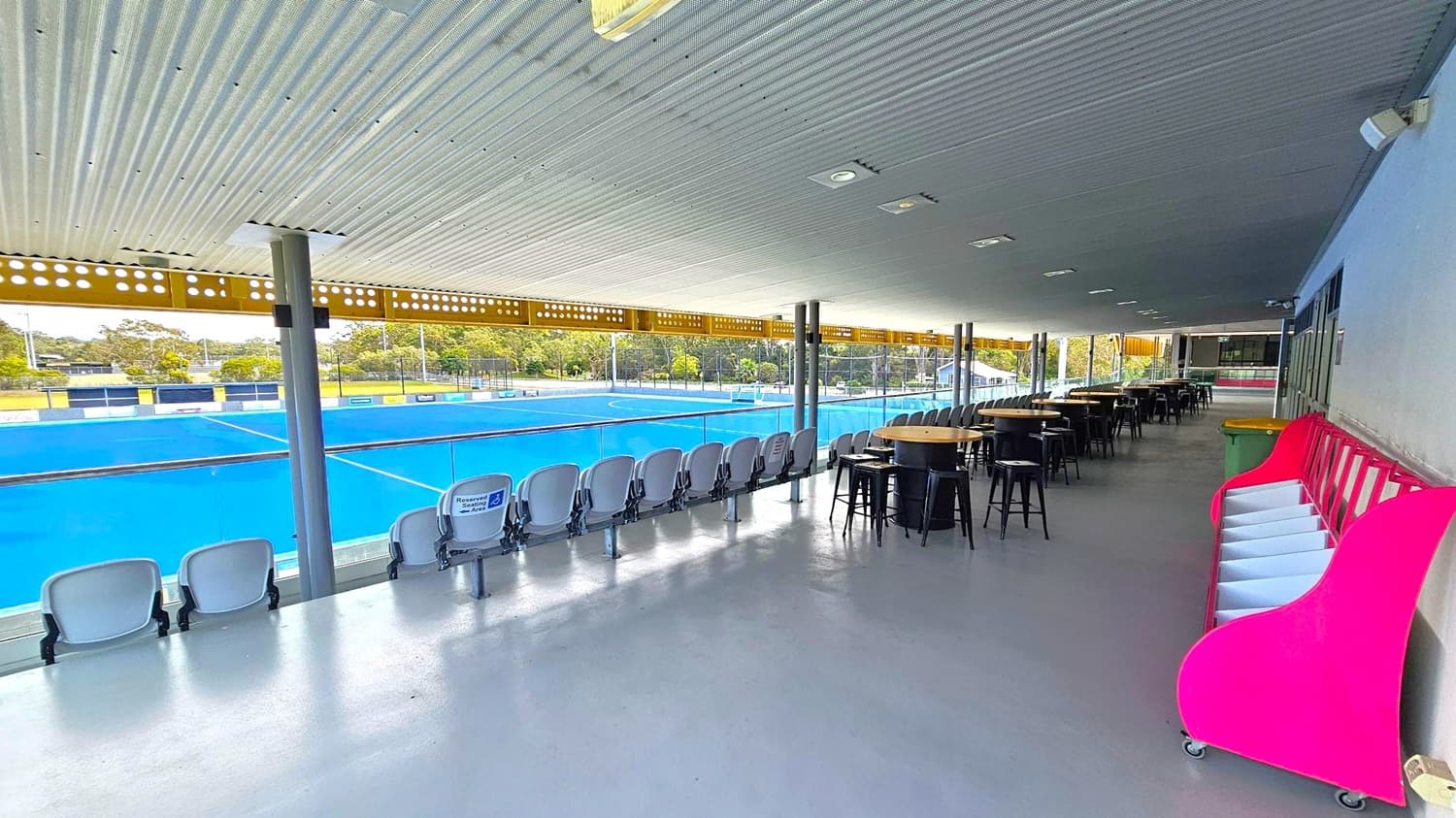 A covered viewing area overlooking a blue sports field. On the left, there are rows of grey seats facing the field. In the middle, several round tables with black stools are arranged. On the right, a bright pink storage unit is against the wall, next to a green bin. The ceiling is corrugated metal, and the floor is smooth concrete. The background shows trees and a fence surrounding the field.