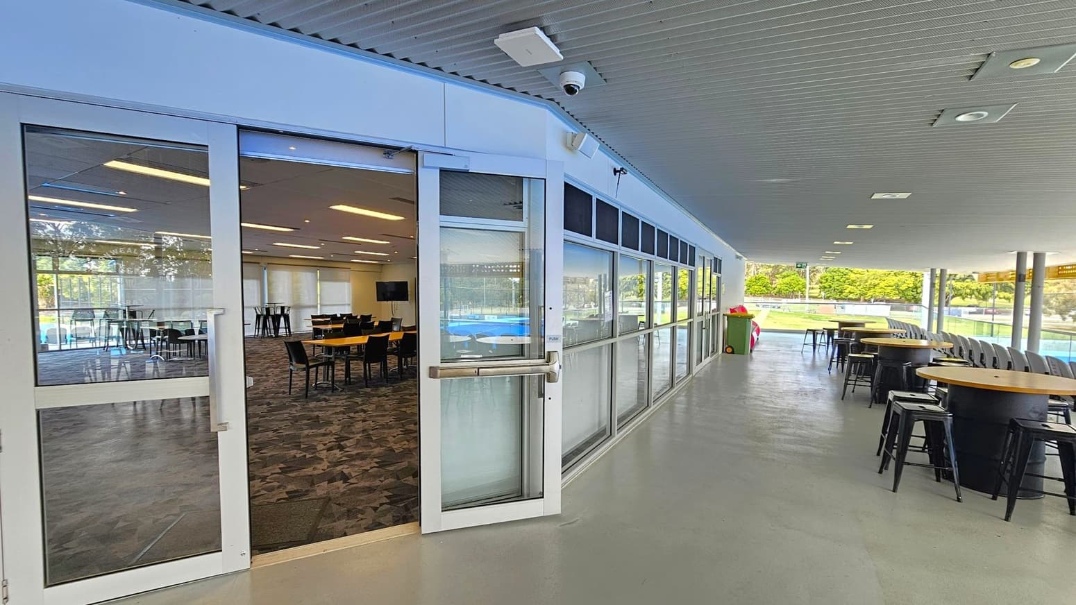 On the left, there is a glass door leading to an indoor area with tables and chairs on a patterned carpet. The right side features an outdoor covered walkway with high tables and stools on a smooth floor. The ceiling is lined with recessed lights. In the background, there is a view of a green outdoor area with trees and a sports field.