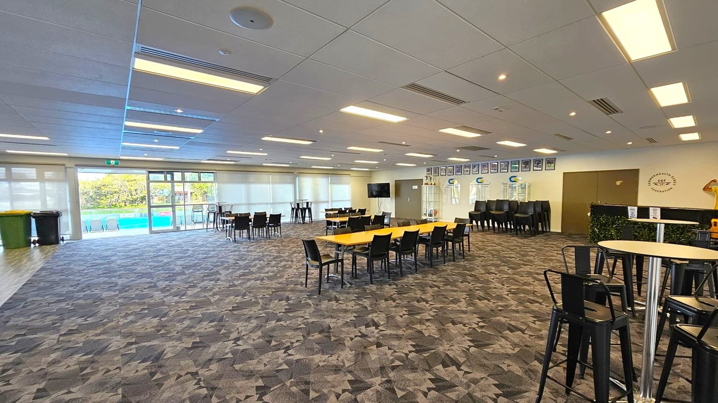 A spacious room with a patterned carpet floor and a ceiling with recessed lighting. On the left, large windows and glass doors lead to an outdoor area with a pool. In the centre, several tables with black chairs are arranged. On the right, a wall displays framed pictures and a logo. There are stacked chairs and a television near the wall. The room has a modern, open feel with ample natural light.