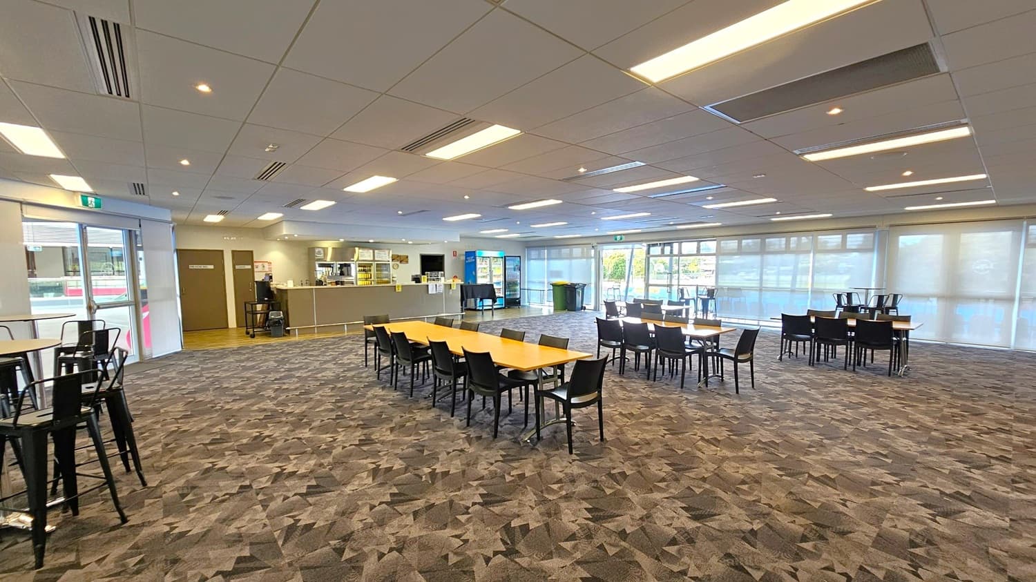 A spacious room with patterned carpet flooring and a grid-like ceiling with recessed lighting. In the centre, there are several tables with black chairs arranged around them. On the left, a bar area with a counter and shelves is visible, along with a door. The right side features large windows with blinds, allowing natural light to enter. In the background, there are more tables and chairs, and a vending machine near the windows.