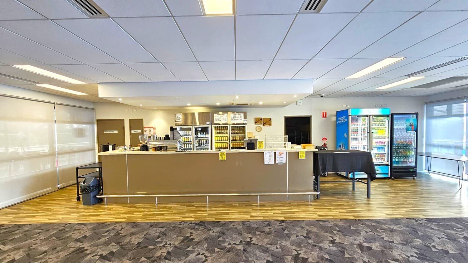 A café counter with a light brown front and a metal footrest runs across the centre. Behind it, shelves and fridges display various drinks and snacks. To the right, two large blue-lit refrigerators are filled with beverages. The floor is a mix of wood and patterned carpet, while the ceiling is lined with white panels and recessed lighting. Blinds cover the windows on both sides, and a small table with a black cloth is near the fridges.