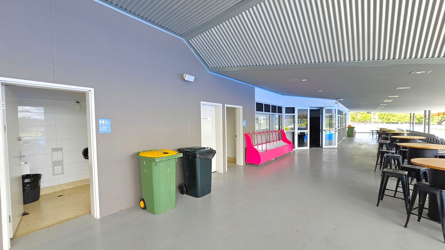 On the left, there is an open door leading to a restroom, with a blue sign indicating accessibility. Next to it are green and black rubbish bins. In the middle, a pink bench is positioned against the wall. On the right, several round wooden tables with black stools are arranged in a row. The ceiling is corrugated, and the floor is smooth and grey. In the background, glass doors and windows lead to an outdoor area with greenery visible.