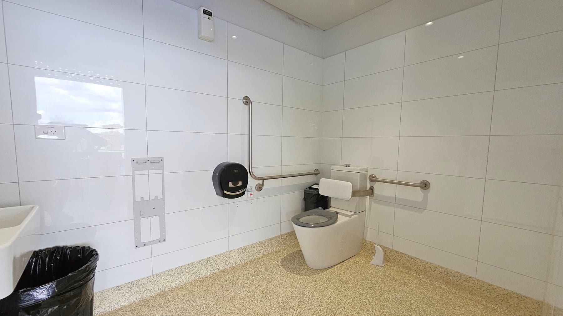 A bathroom with white tiled walls and a speckled beige floor. On the right, there is a toilet with a grey seat, equipped with horizontal and vertical grab bars. Above the toilet is a black toilet paper dispenser. To the left, a black bin is placed under a white sink. An empty wall mount is visible on the left wall, along with an electrical outlet. The ceiling is light-coloured with recessed lighting.