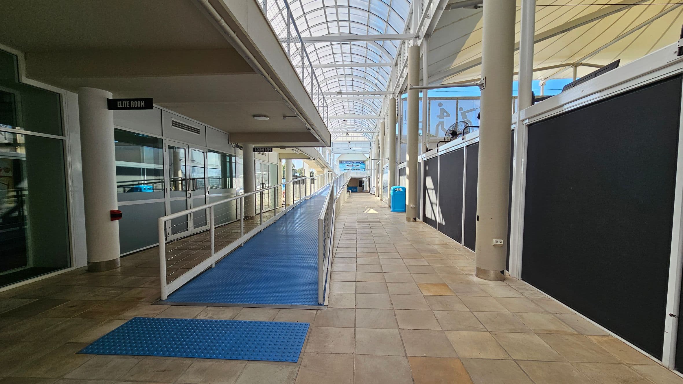 A corridor with a blue ramp on the left, leading to a room labelled "Elite Room". The floor is tiled in beige and light brown. On the right, there are black partition walls and a blue recycling bin. The ceiling is a transparent, arched structure allowing natural light. A beige column supports the ceiling on the right side.