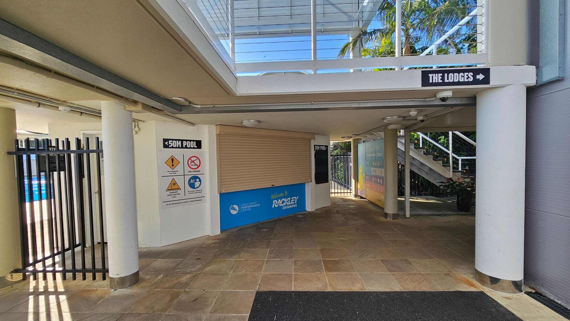 A covered walkway with tiled flooring. On the left, there's a black metal gate leading to a pool area, with signs indicating pool rules. In the centre, a closed shutter with a sign for "Rackley Swimming" is visible. On the right, a sign points to "The Lodges" with a staircase leading upwards. The ceiling is supported by white columns, and there's a glass section above, allowing natural light in. Palm trees are visible through the glass.