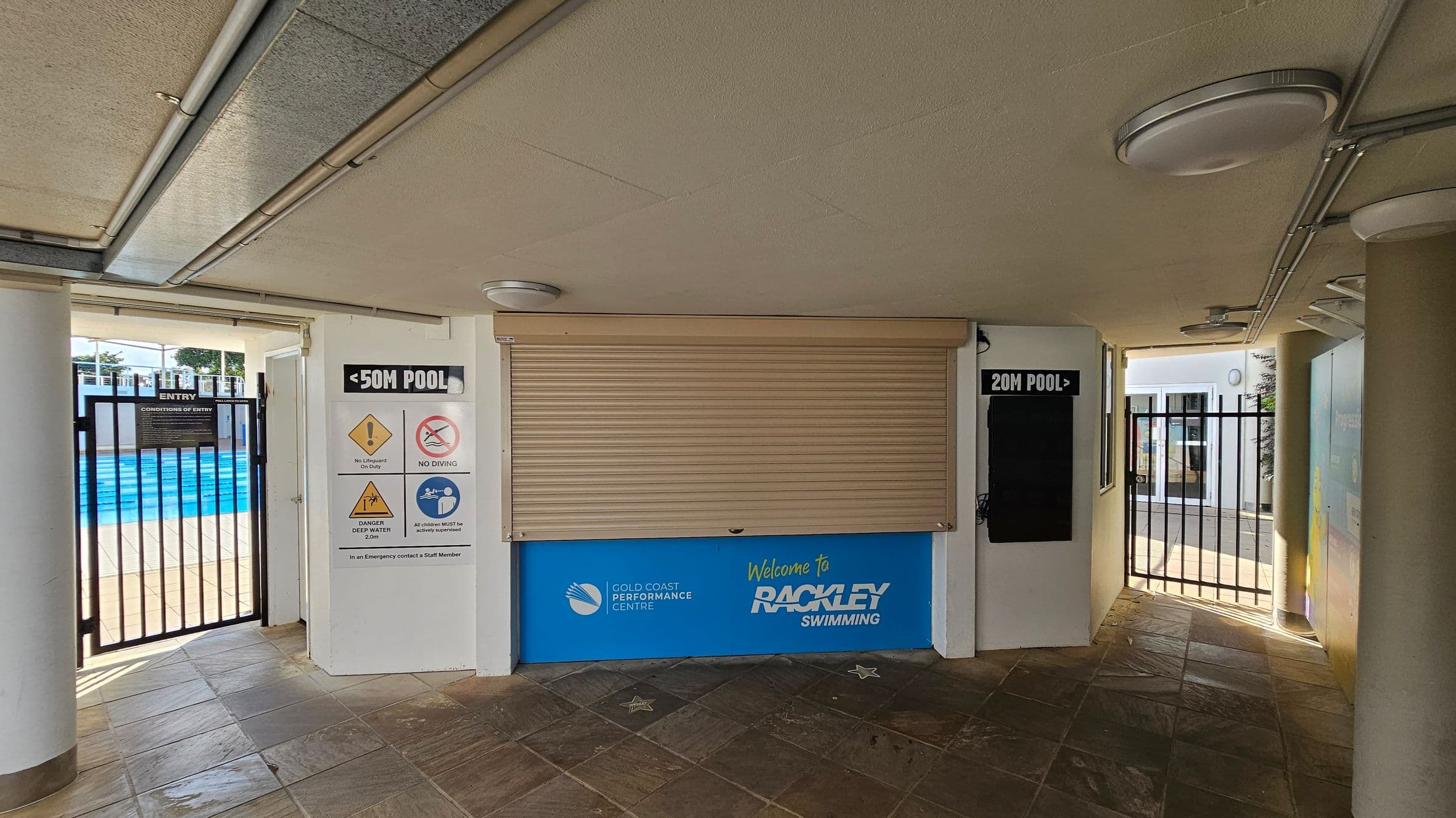 A corridor with a tiled floor and a ceiling with circular lights. In the centre, a closed roller shutter with a blue sign below reading "Welcome to Rackley Swimming" and "Gold Coast Performance Centre." On the left, a gate with signs indicating a 50m pool and safety warnings. On the right, another gate leading to a 20m pool. Both gates have black bars and are open to outdoor areas.