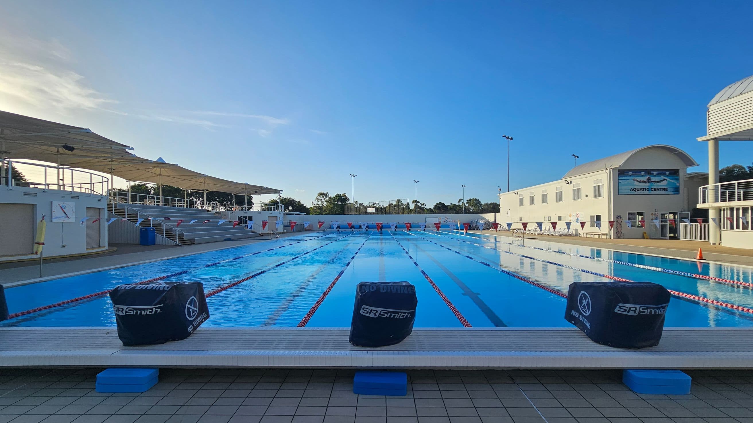 An outdoor swimming pool with multiple lanes, bordered by a tiled deck. On the left, there are shaded seating areas with white canopies and steps leading up. On the right, a white building with a sign reading "Aquatic Centre" and several windows. The pool is divided by lane ropes, and there are "No Diving" signs on the starting blocks. In the background, trees and tall lights are visible.