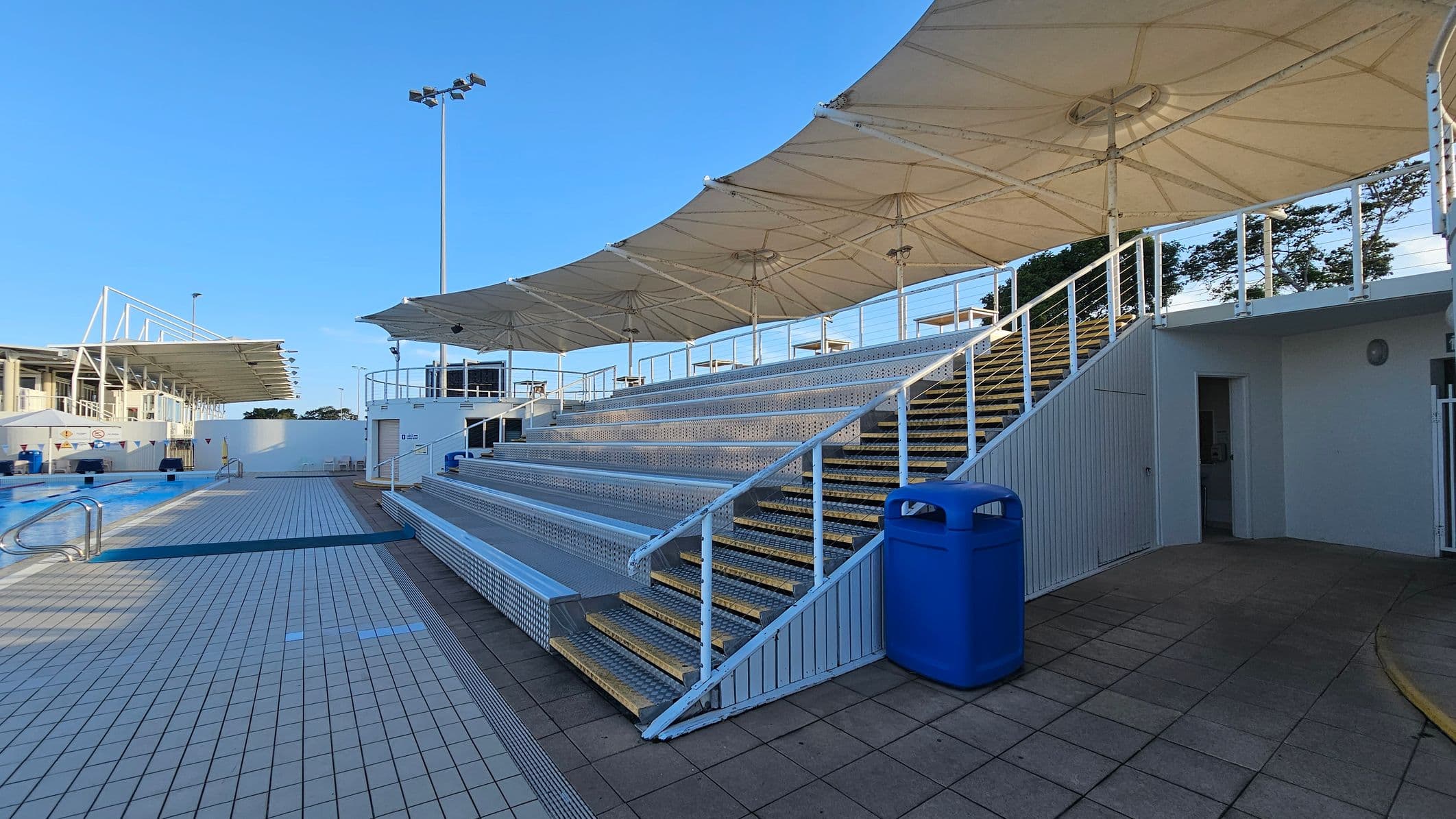 Outdoor swimming pool area with a tiled deck on the left and a set of metal bleachers on the right. The bleachers are covered by large, white, umbrella-like canopies. A blue rubbish bin is positioned near the stairs leading up the bleachers. In the background, there are tall floodlights and a building with a similar canopy structure. The floor is a mix of light and dark tiles, and there is a small open doorway under the bleachers.