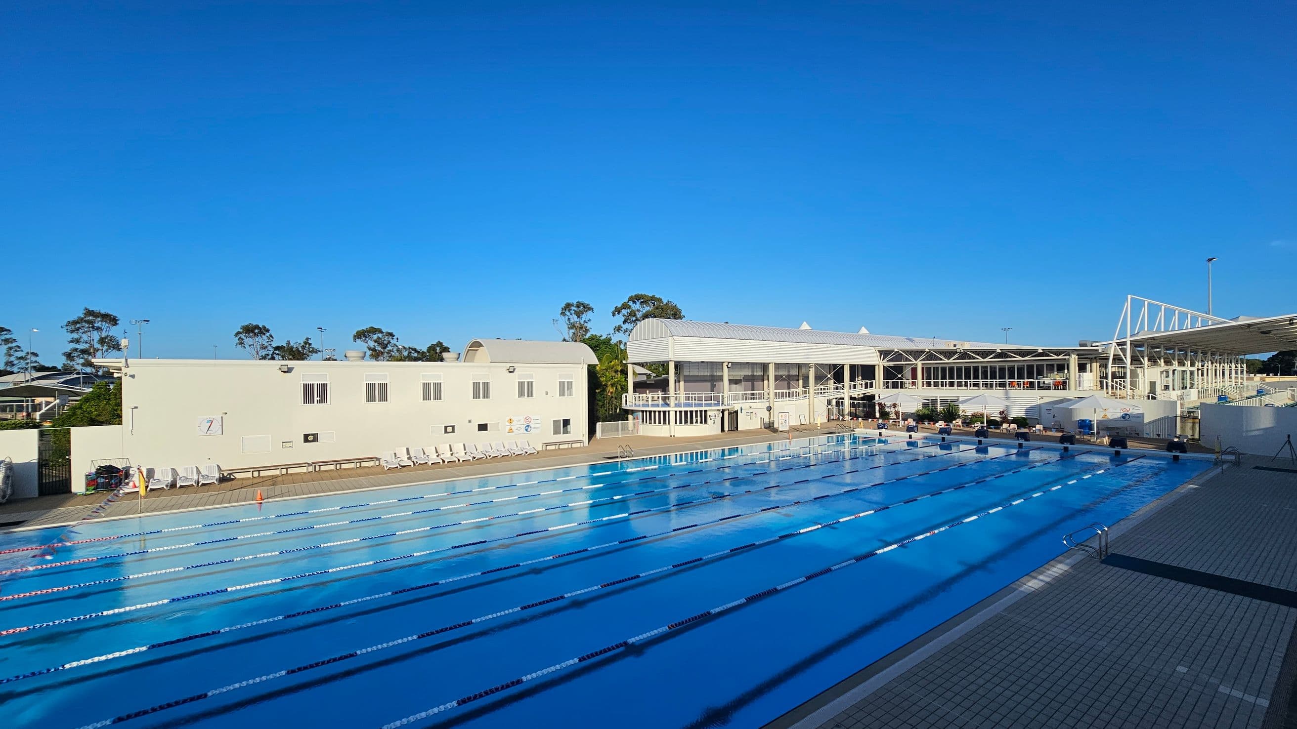 An outdoor swimming pool with multiple lanes stretches across the foreground, bordered by a tiled deck. On the left, a light-coloured building with a curved roof and several windows is visible, with white deck chairs lined up in front. In the centre and right, a larger building with a modern design features glass windows and a white framework. Trees are visible in the background, and the sky is clear.