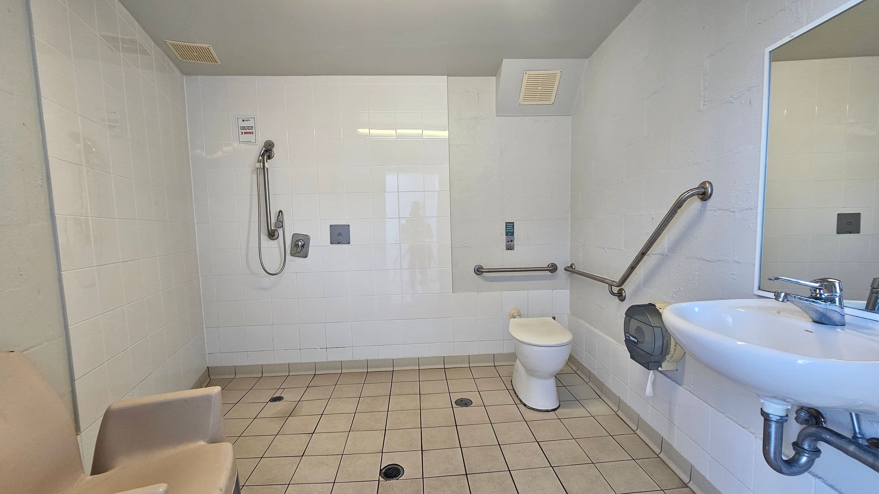 A spacious, accessible bathroom with light-coloured tiled walls and floor. On the left, there's a shower area with a handheld showerhead and a wall-mounted seat. In the centre, a toilet with grab bars on both sides. On the right, a sink with a mirror above it and a paper towel dispenser nearby. The ceiling is plain with a vent. The room is designed for accessibility and ease of use.