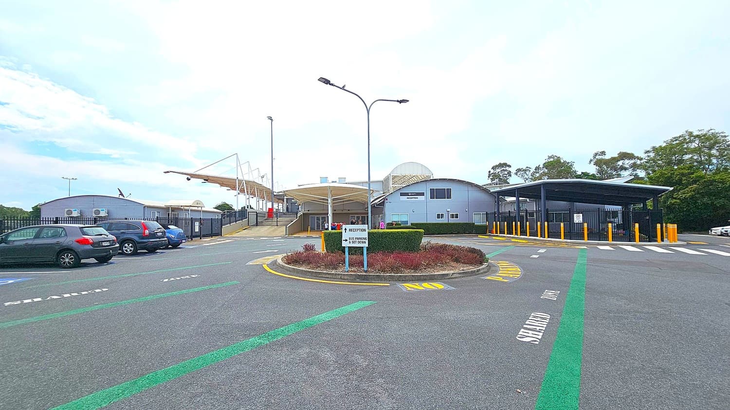 A parking area with several cars on the left. In the centre, a roundabout with a sign pointing to "Reception" and "Bus Parking & Set Down". The building in the background has a curved roof and a "Reception" sign. On the right, a covered area with yellow bollards and a pedestrian crossing. The ground has marked lanes, and there are trees in the background.