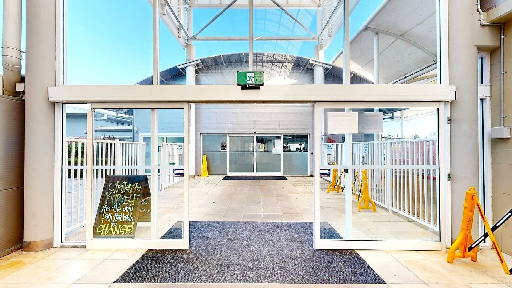A spacious entrance with large glass sliding doors and a high ceiling. In the foreground, a dark mat lies on a light-coloured tiled floor. A sign on the left reads, "Challenge yourself, it's the only path that leads to change!" Beyond the doors, a corridor with more glass and metal structures is visible, along with yellow caution signs. The ceiling features a metal framework with a green emergency exit sign above the doors.