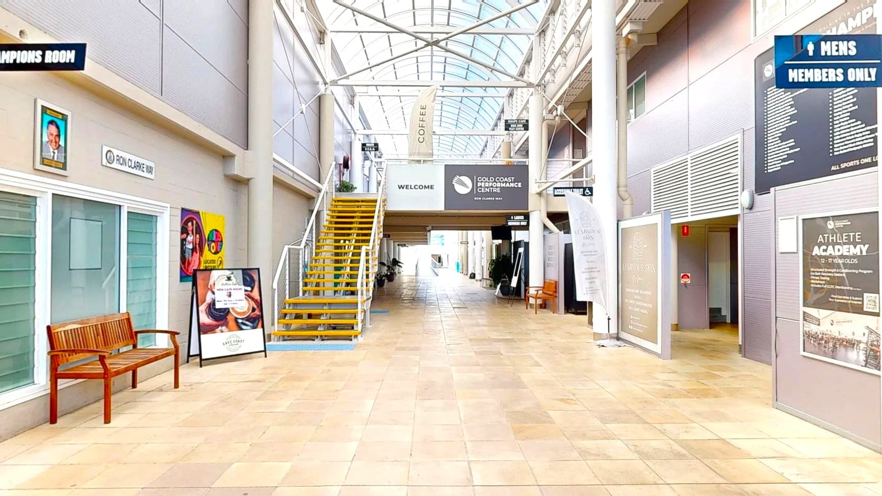 A spacious indoor corridor with a high, arched glass ceiling. On the left, there's a wooden bench in front of a glass-fronted room with a sign above it. A staircase with yellow steps leads to an upper level. On the right, various signs and posters are displayed on the walls, including one for an athlete academy. The floor is tiled in a light beige colour, and the corridor extends into the distance with more signs and seating visible.