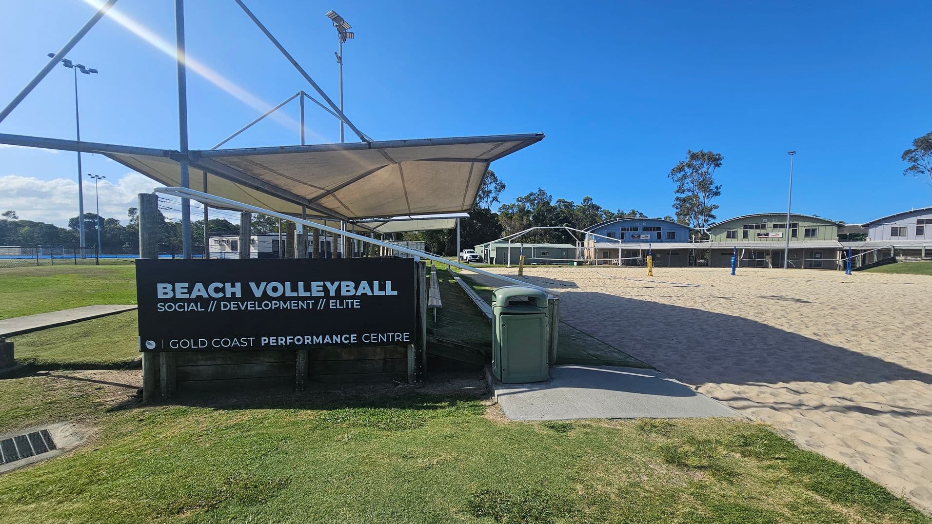 On the left, a black signboard with white text reads "Beach Volleyball Social // Development // Elite" above "Gold Coast Performance Centre," positioned near a grassy area with a nearby concrete drain cover. In the middle, a large fabric shade structure supported by metal poles covers a walkway. On the right, sandy volleyball courts with yellow and blue poles extend toward three green and grey rectangular buildings in the background. The floor alternates between grass, sand, and pavement.