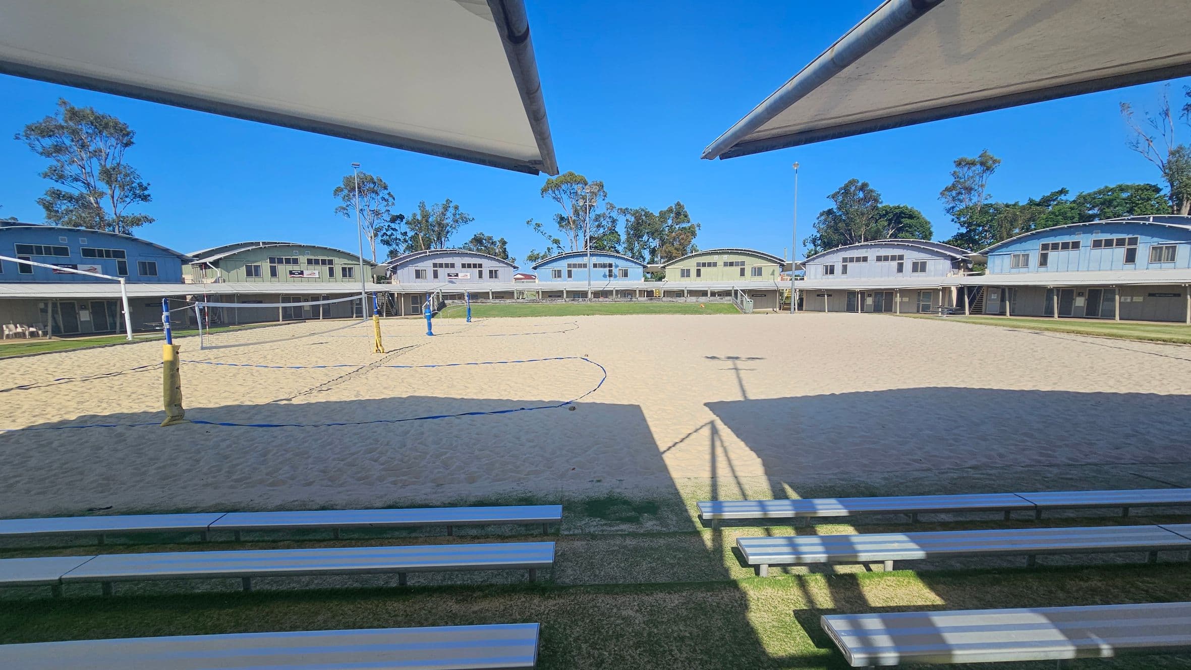 On the left, white shelters extend over the edges of a sandy volleyball court marked with blue boundary lines and yellow poles holding up nets. In the middle, multiple single-storey rectangular buildings with arched roofs and large windows form the background. On the right, more stadium-style seating made of metallic benches is visible along the grassy border. The foreground has tiered metal benches, while the ceiling consists of the edges of fabric sunshades.