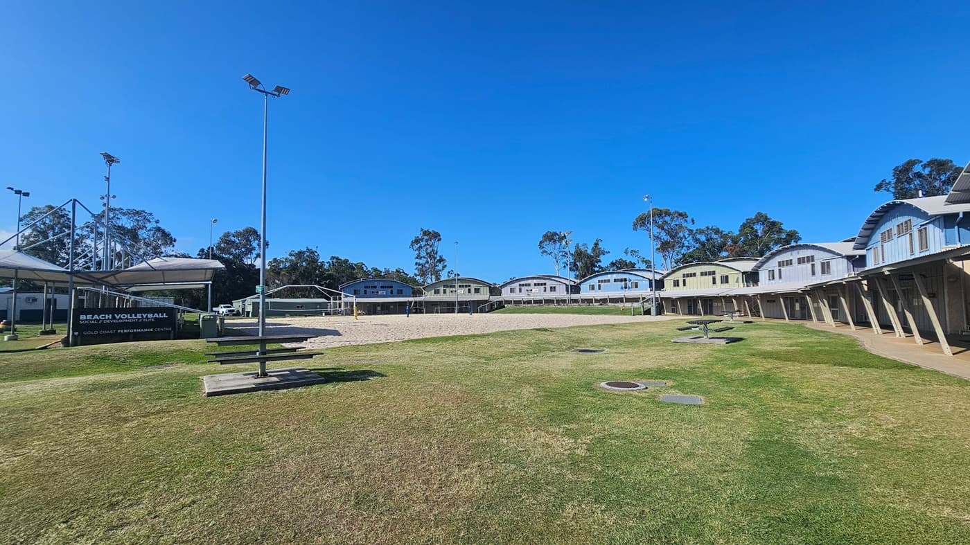 On the left, a black sign with white text reading "Beach Volleyball Gold Coast Performance Centre" sits near a shaded seating structure with metal framing. In the middle, a sandy volleyball court is enclosed by yellow posts and surrounded by a row of pale green and white arched-roof buildings. On the right, more buildings with covered walkways line a grassy area with picnic tables and drainage covers. The ground alternates between grass, sand, and pavement.