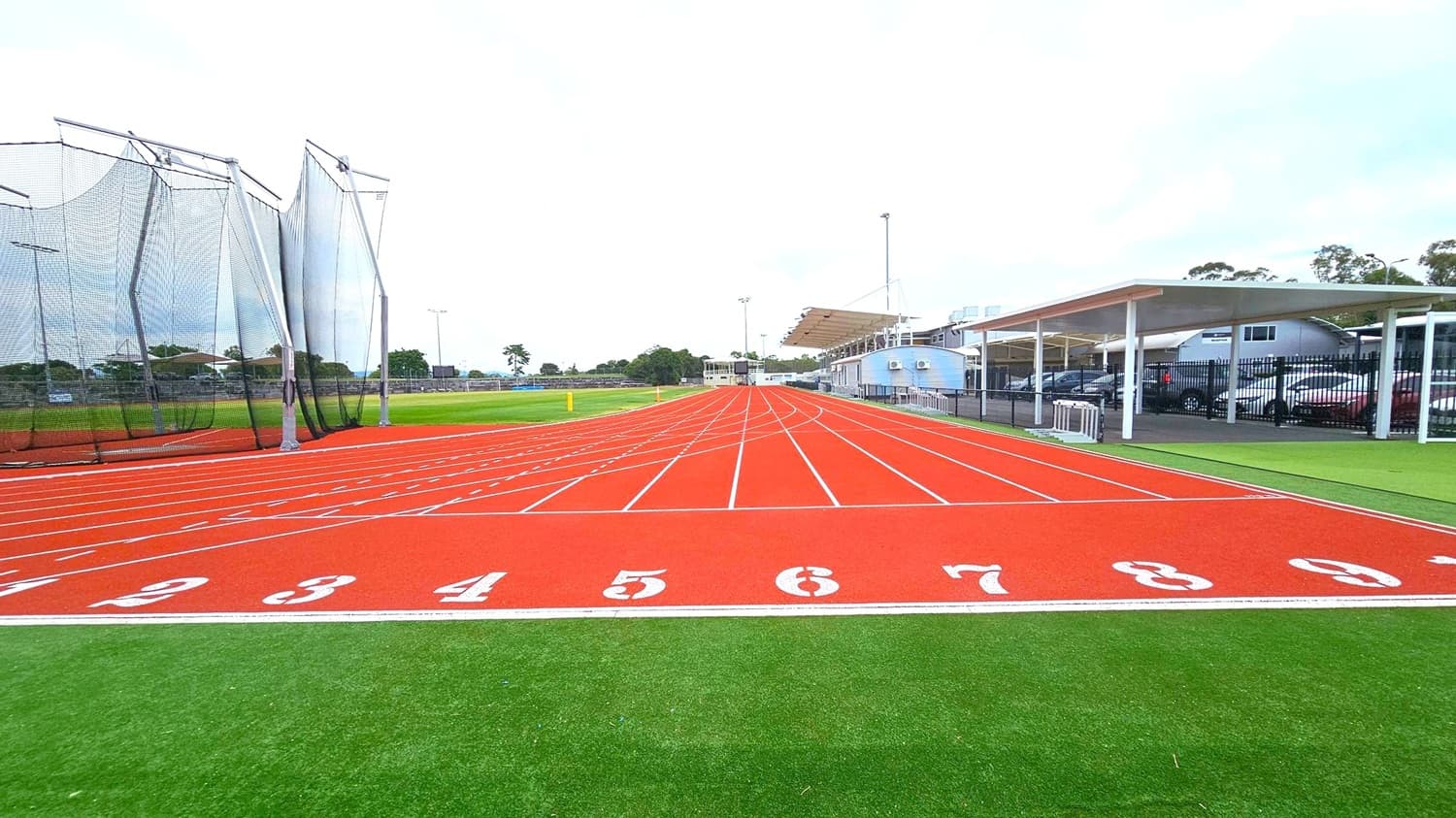 A red athletics track with white lane numbers from 1 to 9 in the foreground. On the left, there are tall netted structures, possibly for field events. To the right, a covered area with parked cars and a building with a sloped roof. The background features more buildings and trees. The track is surrounded by green artificial turf.