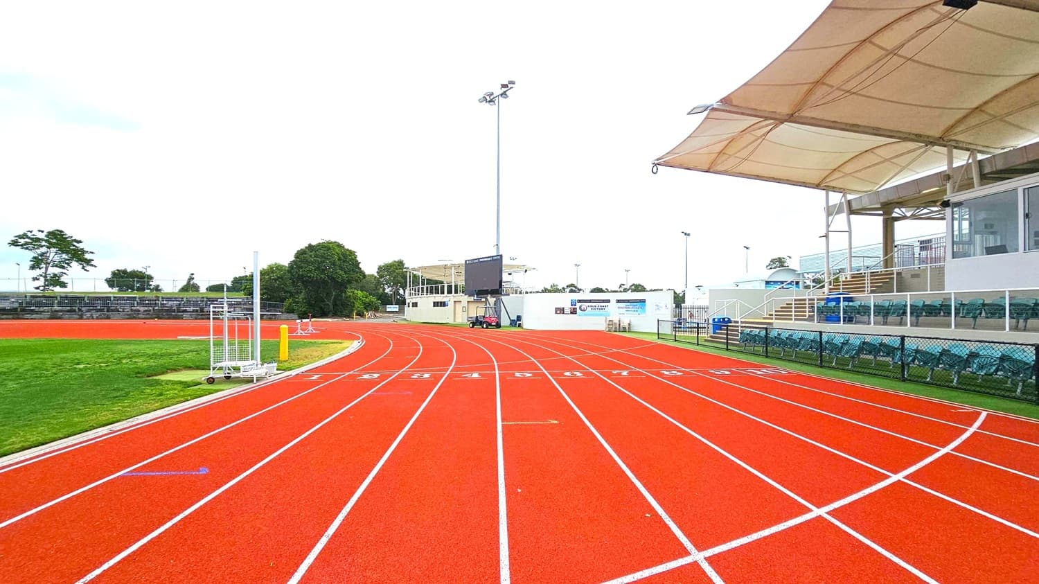 A red athletics track with white lane markings curves to the right. On the left, there's a grassy area with some equipment and trees in the background. On the right, a covered seating area with green chairs is visible, along with a white structure and stairs. A large screen and light poles are in the distance. The area is enclosed by a low fence.