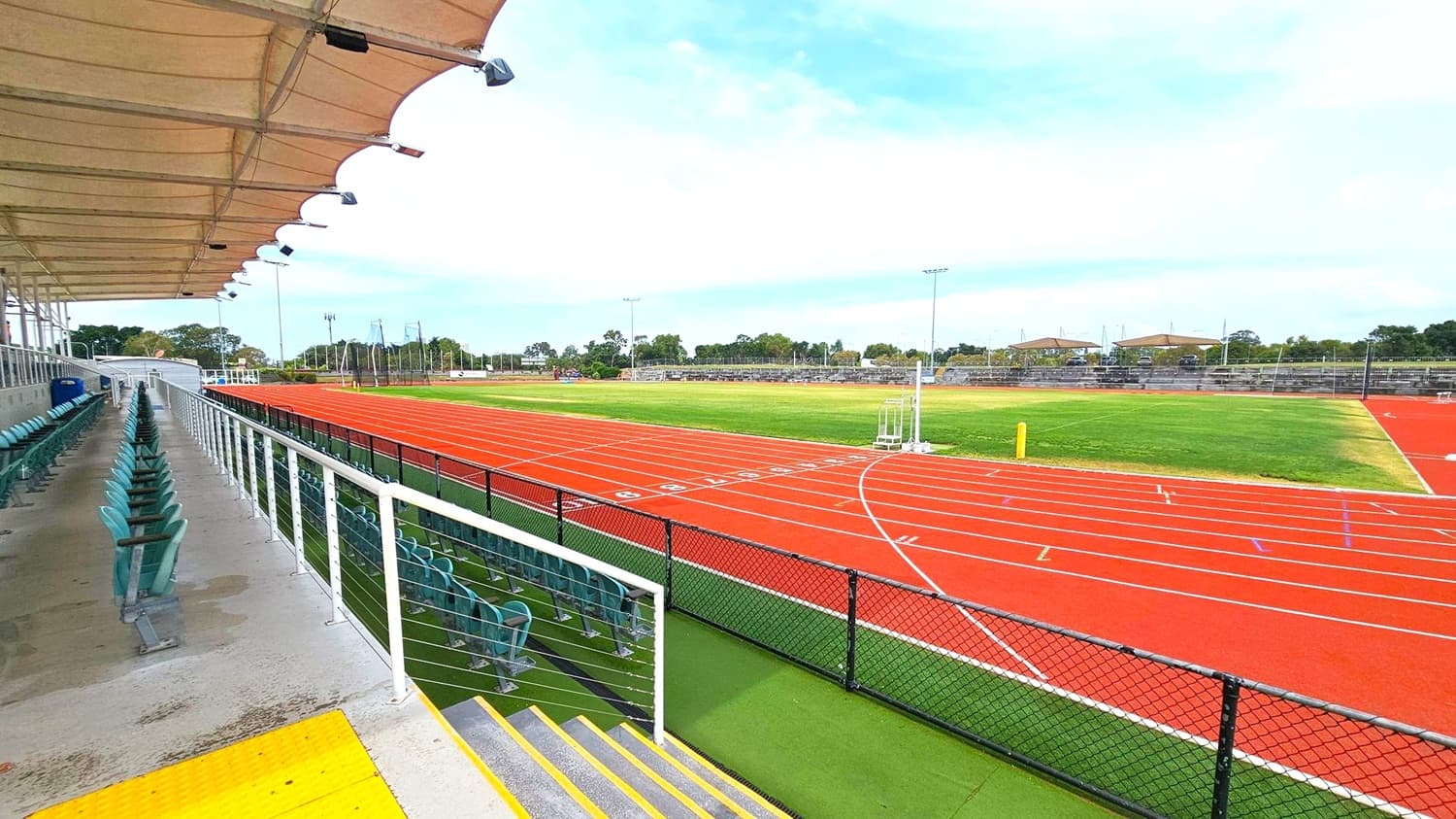 On the left, there is a covered seating area with rows of green chairs and a white railing. The ceiling is a fabric canopy with lights attached. The foreground features a yellow tactile paving strip at the top of the steps. On the right, a red athletics track with white lane markings encircles a green grass field. In the background, there are more seating areas and trees, with several light poles around the track.