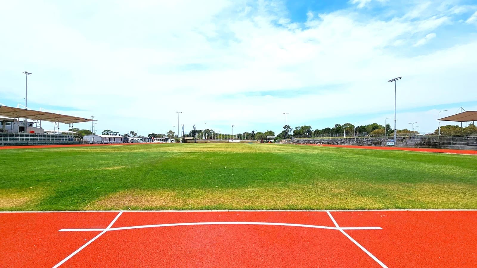 A sports field with a red running track in the foreground, marked with white lines. The grass in the middle is a mix of green and brown patches. On the left and right, there are covered spectator stands with metal railings. Several tall floodlights are positioned around the field. In the background, there are trees and some small buildings.
