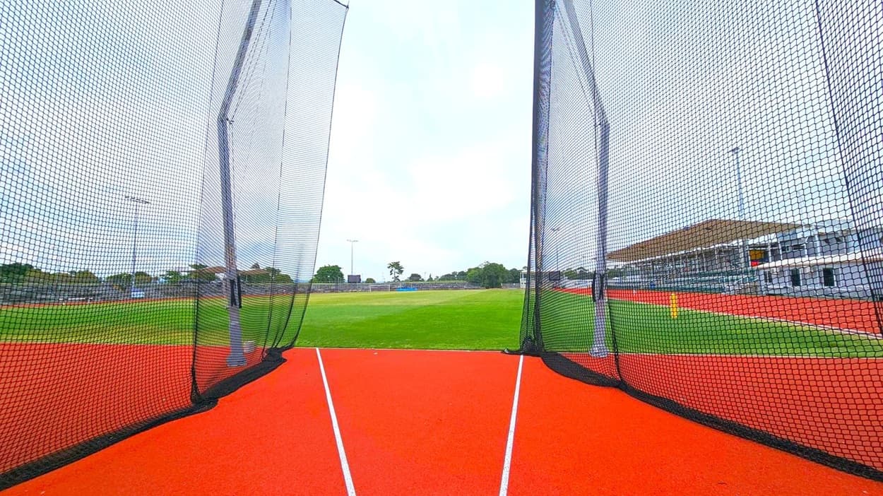 A sports field with a red running track in the foreground, bordered by black netting on both sides. The netting is supported by tall poles. Beyond the track, a green grass field extends into the distance. On the right, there is a large building with a sloped roof, possibly a stadium or sports facility. Trees and additional structures are visible in the background, along with several floodlights.