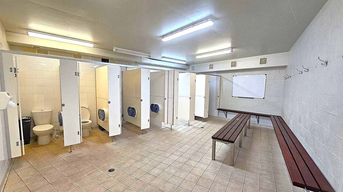 A changing room with a row of toilet cubicles on the left, each with a white door and a toilet inside. The floor is tiled in beige and grey. On the right, there are wooden benches and a series of hooks on the wall. A whiteboard is mounted on the wall above the benches. The ceiling is fitted with fluorescent lights, providing bright illumination.