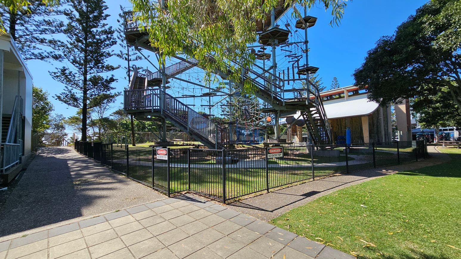 A high ropes course with multiple platforms and obstacles is enclosed by a black metal fence with "Danger No Entry" signs. On the left, a gravel path runs alongside a building, leading towards tall trees. On the right, a grassy area and a large tree provide shade. In the background, a structure with a curved roof is visible. The ground is a mix of paved tiles and grass.