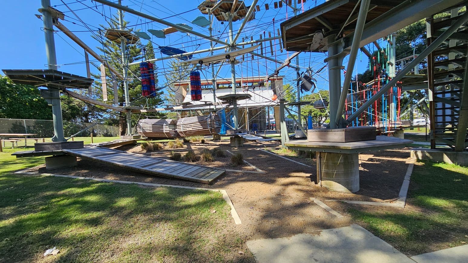 An outdoor adventure park with a high ropes course. The structure is made of metal poles and wooden platforms connected by ropes and cables. There are various obstacles, including hanging logs, rope bridges, and vertical climbing elements. The ground is a mix of grass and dirt, with some patches of sunlight and shadow. In the background, there are trees and a fence.