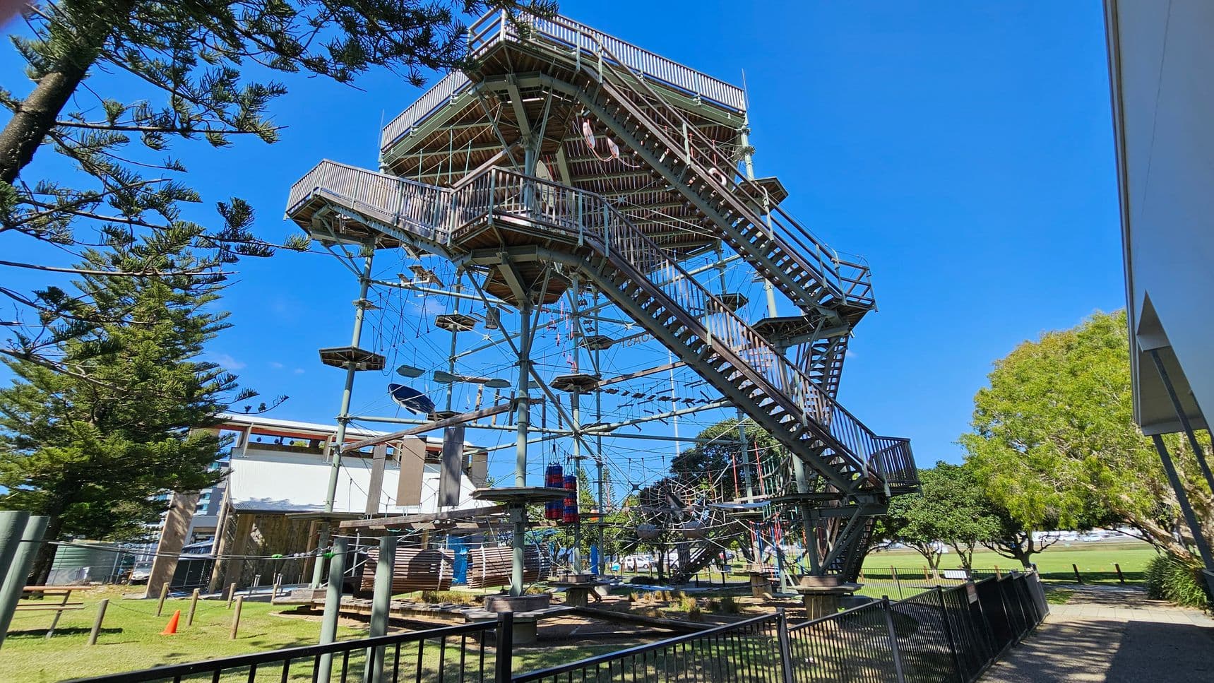 A large outdoor climbing structure with multiple levels and platforms, featuring ropes and obstacles. It is surrounded by a metal fence. On the left, there are tall trees with dense foliage. On the right, a building with a white wall is partially visible, and more trees are in the background. The ground is grassy, and a bright orange cone is placed near the structure.