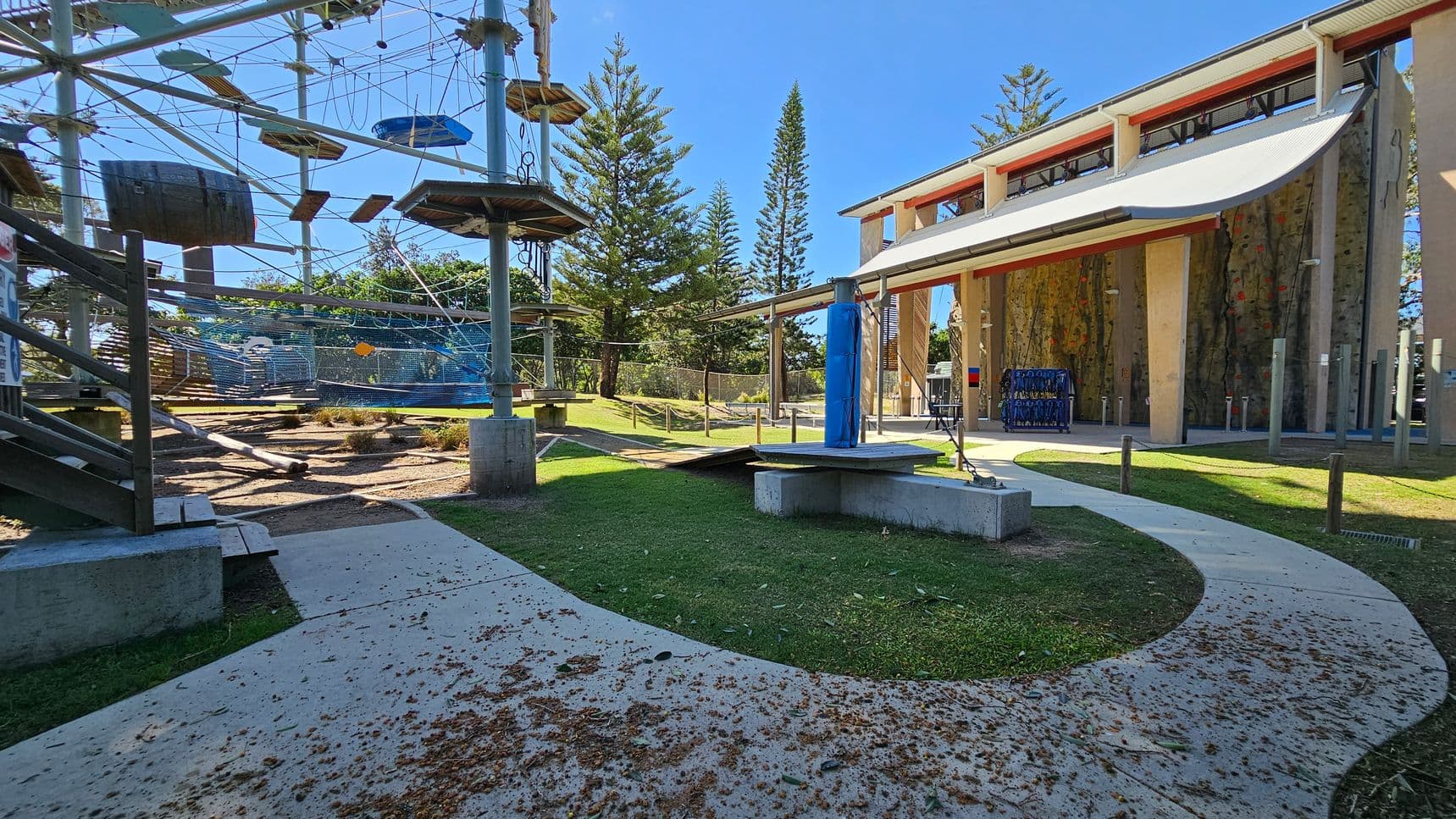 An outdoor adventure park with a high ropes course on the left, featuring platforms, ropes, and a barrel. In the centre, a curved concrete path with scattered leaves leads to a grassy area. On the right, a building with a large canopy and climbing walls. Tall trees are visible in the background.