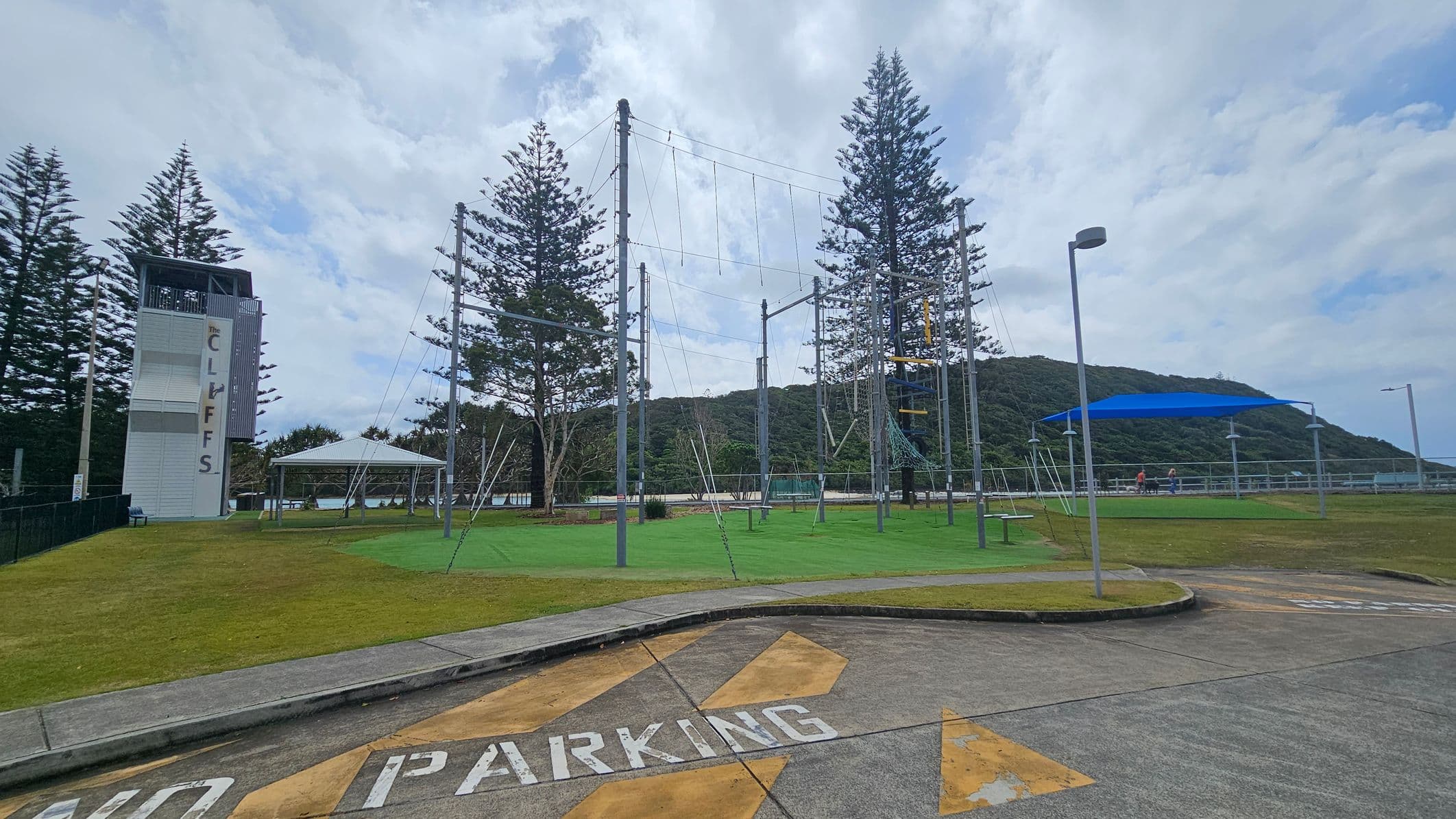 A concrete area with "NO PARKING" painted on it is in the foreground. To the left, there's a tall structure labelled "THE CLIFFS" next to a grassy area with a white canopy. In the centre, a high ropes course with metal poles and wires is visible. To the right, a blue shade structure covers a section of the grass, with a few people nearby. Tall trees and a hill are in the background.