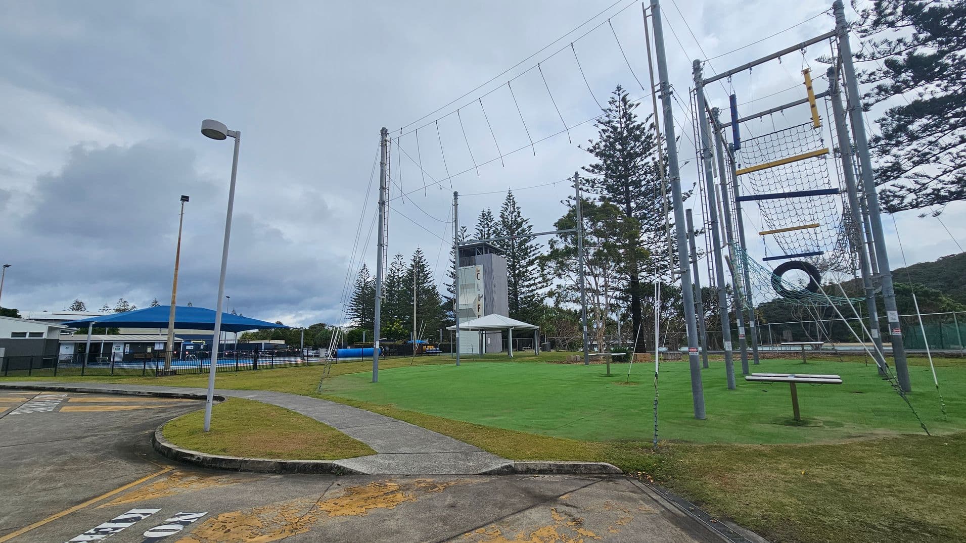 A recreational area with a high ropes course on the right, featuring nets, ropes, and a tyre. In the centre, there's a tall climbing tower with a small shelter nearby. On the left, a building with a blue roof is visible, surrounded by a black fence. The ground is a mix of concrete paths and green grass, with a few scattered trees in the background. A streetlamp stands in the foreground.