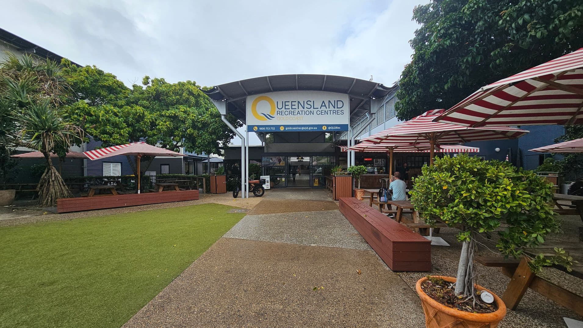 A courtyard with a pathway leading to the entrance of the Queensland Recreation Centres. The building has a large sign above the entrance. On the left, there are trees and picnic tables under red and white striped umbrellas. On the right, more picnic tables and benches are shaded by similar umbrellas. A potted tree is in the foreground on the right. The ground is a mix of grass and paved areas.