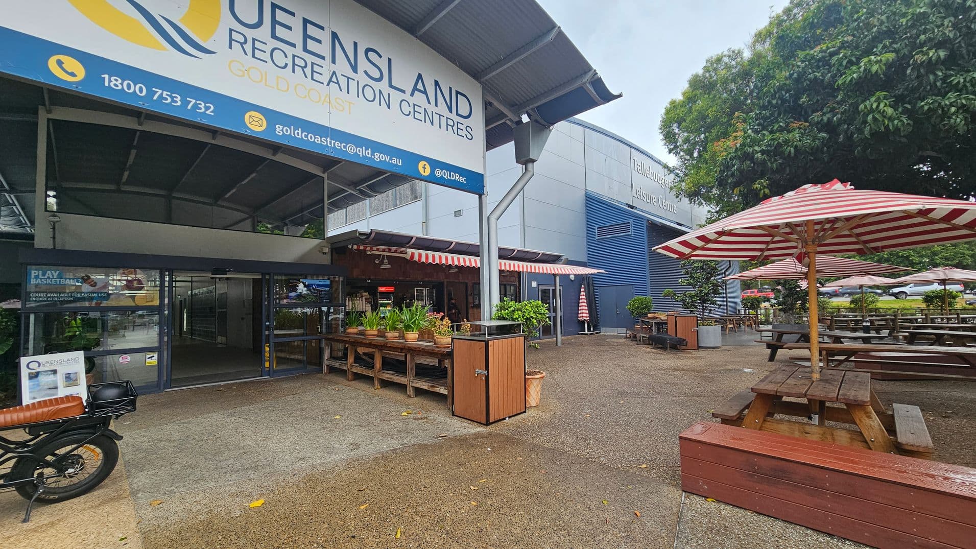 Entrance to Queensland Recreation Centres Gold Coast with a large sign above. On the left, a motorbike is parked near the entrance. In the centre, there are potted plants on wooden tables under a canopy with red and white striped awnings. On the right, picnic tables with matching striped umbrellas are set on a paved area. A large tree provides shade in the background, and a building labelled "Tallebudgera Leisure Centre" is visible.