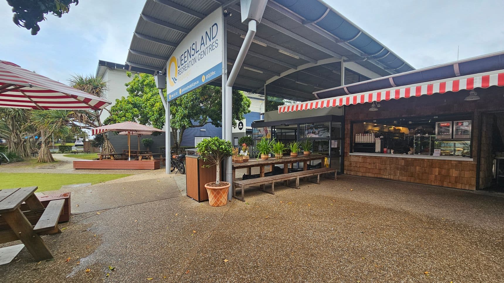 Outdoor seating area with wooden picnic tables and benches on the left, under red and white striped umbrellas. In the centre, a sign reads "Queensland Recreation Centres" above a row of potted plants on a wooden table. On the right, a café with a red and white striped awning displays a counter with food and drinks inside. The ground is a mix of grass and textured pavement, and there are trees in the background.