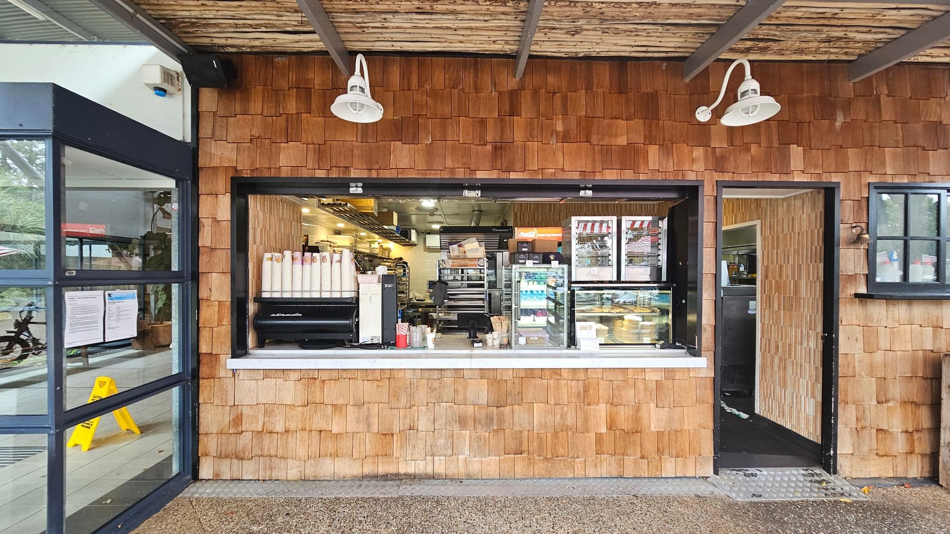 A small café with a wooden shingle exterior. In the centre, there's a service window displaying coffee cups, a coffee machine, and a glass case with pastries. To the right, an open door leads inside. Above, two white lamps are mounted on the wall. On the left, a glass door with notices and a yellow caution sign is visible. The ceiling is made of wooden slats, and the floor is a textured concrete surface.