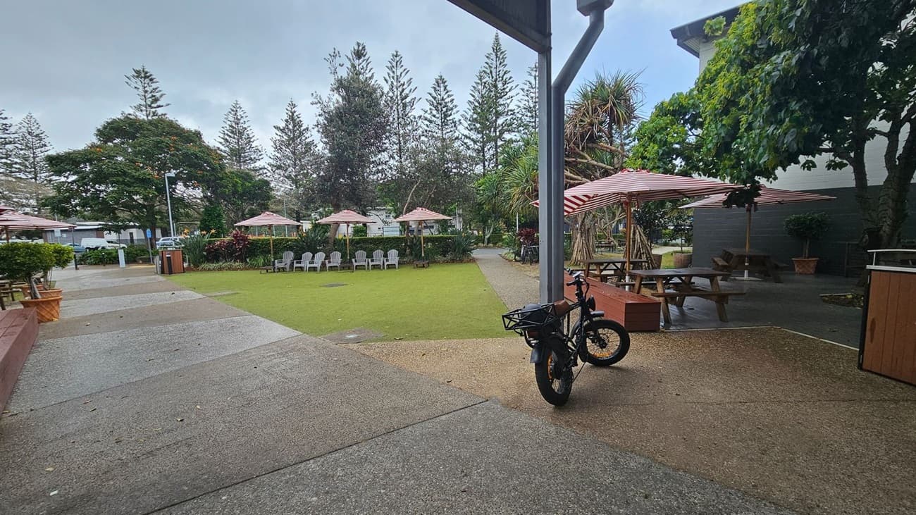 A courtyard with a mix of concrete and grass surfaces. On the left, there are several picnic tables with red and white striped umbrellas. In the centre, a grassy area is surrounded by white chairs and more umbrellas. On the right, a bicycle is parked near a wooden bench. Trees and shrubs are scattered throughout, providing shade and greenery. A building with a dark wall is partially visible on the right.