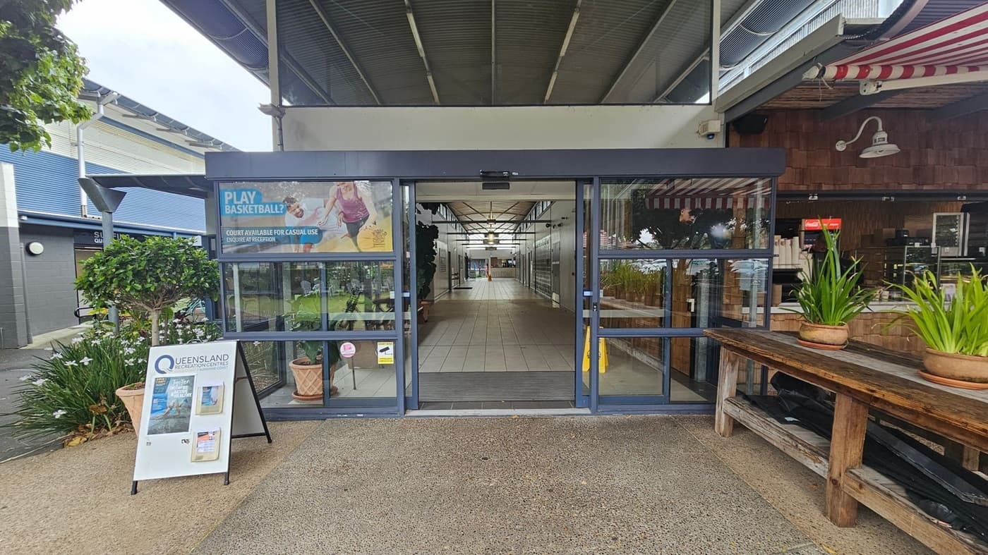 Entrance to a building with glass doors in the centre. A sign on the left reads "Queensland Recreation Centres" with promotional material. A small tree and flowers are nearby. On the right, a wooden bench holds potted plants, and a café with a red and white striped awning is visible. The floor is tiled, and the ceiling has exposed beams and lights.