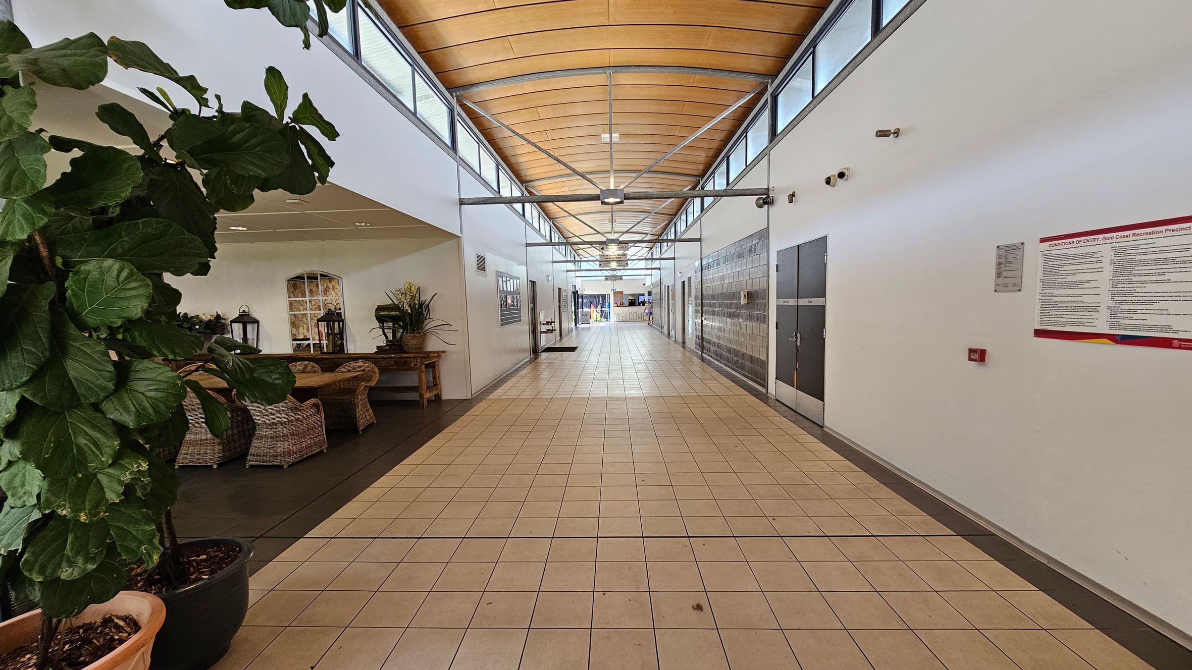 A corridor with a tiled floor and a wooden panelled ceiling. On the left, there are large potted plants and wicker furniture, including chairs and a table with decorative items. The right side features a white wall with a notice board and security cameras. The corridor extends into the distance, with doors and windows along the sides, leading to a bright area at the end.